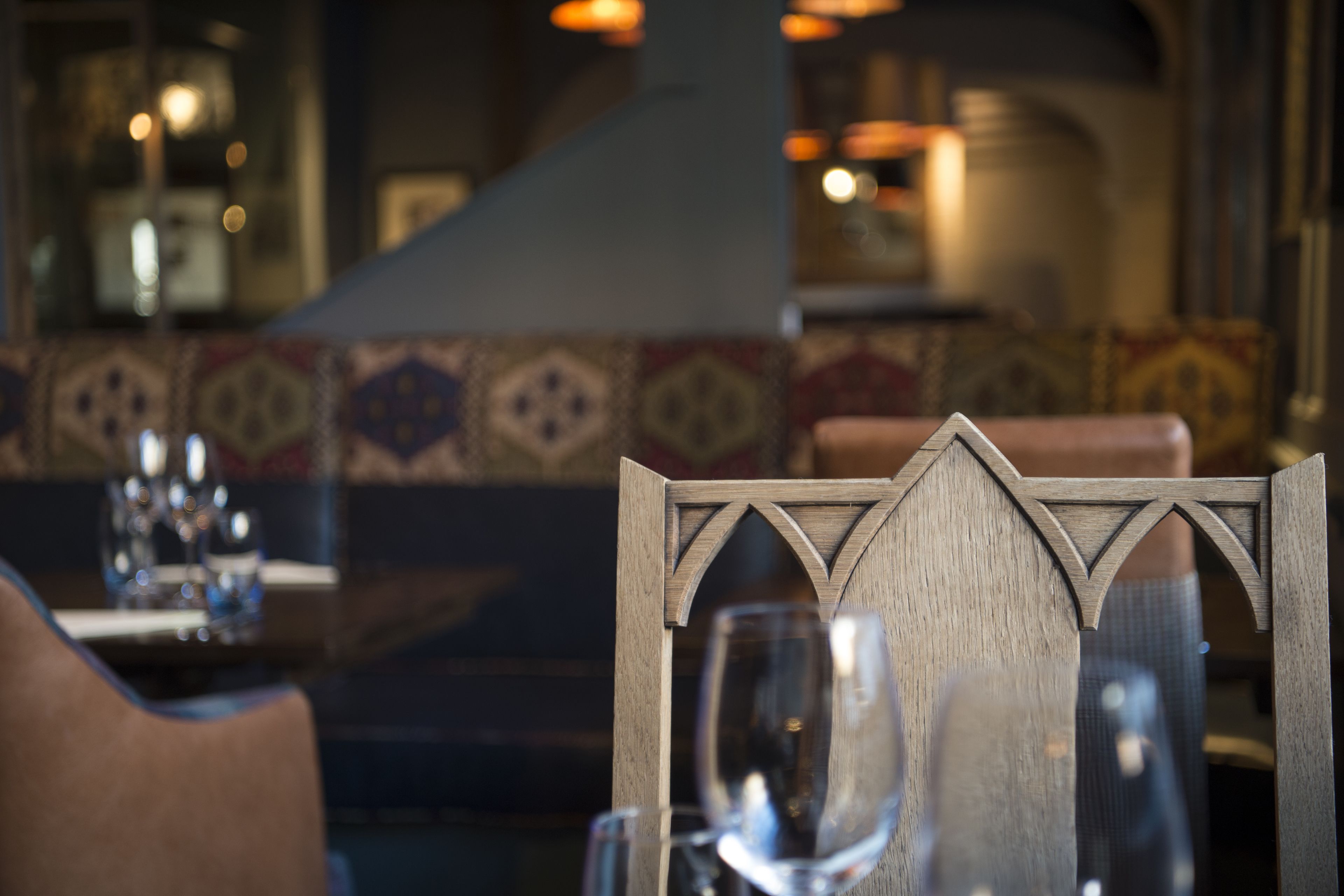 A wooden chair with a geometric backrest design, a glass table with a wine glass, and a patterned wall in the background.