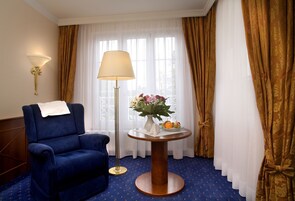Un sillón azul, una mesa redonda de madera con un jarrón de flores y fruta, una lámpara de pie y una ventana con cortinas blancas translúcidas.