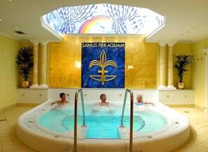 A pool area with three people in the water, a large mural with the text "SANUS PER AQUAM" and a fleur-de-lis symbol, and a skylight above. - Hotel & SPA Fontenay (Bad Woerishofen)