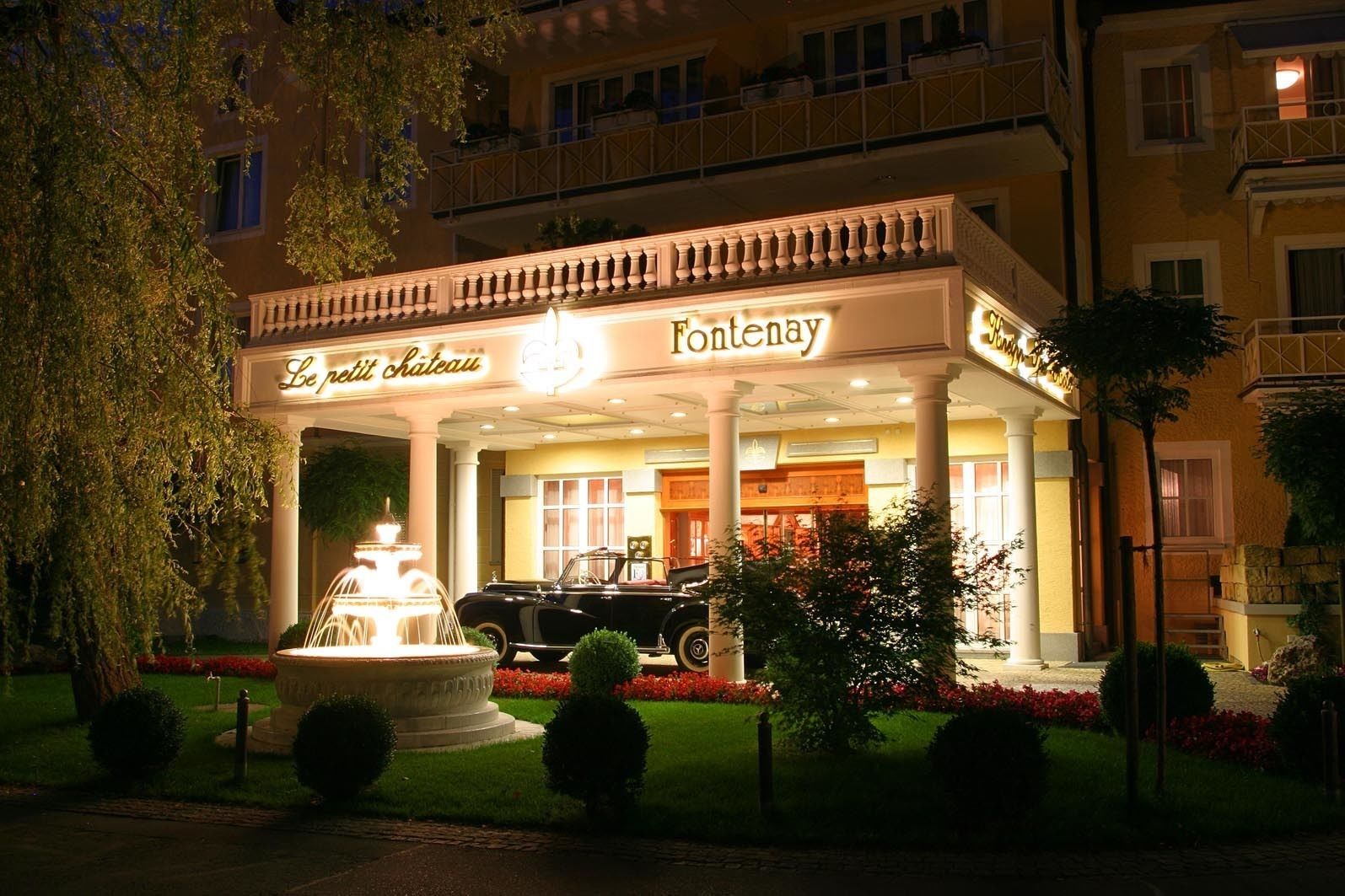 A hotel entrance with a fountain, a classic car, and a sign reading "Fontenay Le petit château."