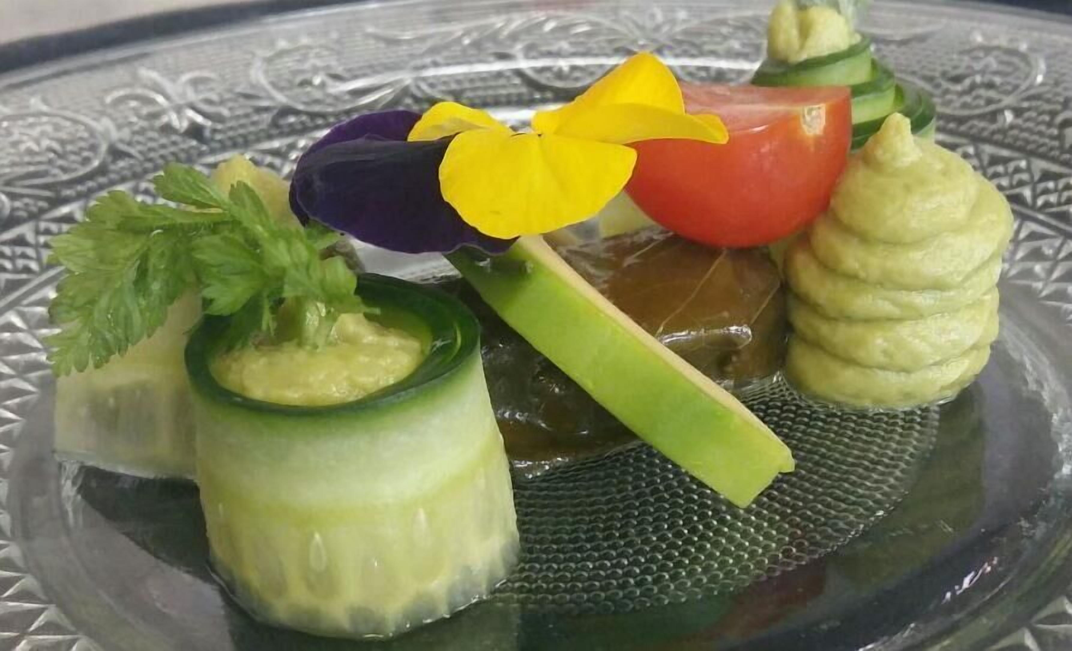 A glass plate with a decorative pattern, containing a variety of food items including a cucumber with a yellow filling, a tomato, a purple vegetable, and a green vegetable with a yellow filling.