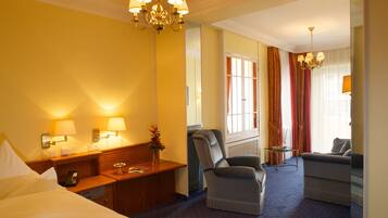 A hotel room with a bed, a desk with a lamp, a chair, a sofa, and a chandelier.