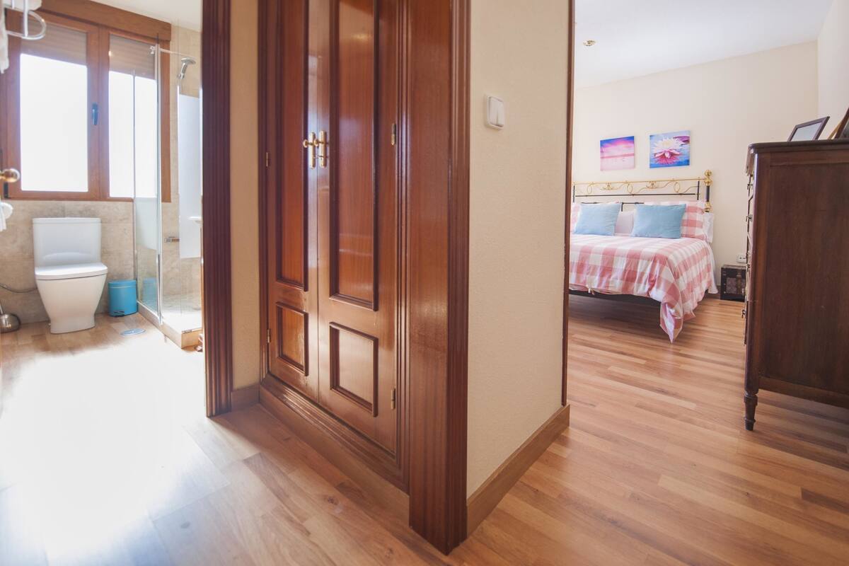 Habitación de hotel con piso de madera, una cama con colcha a cuadros, armario de madera y un baño con inodoro y ducha.