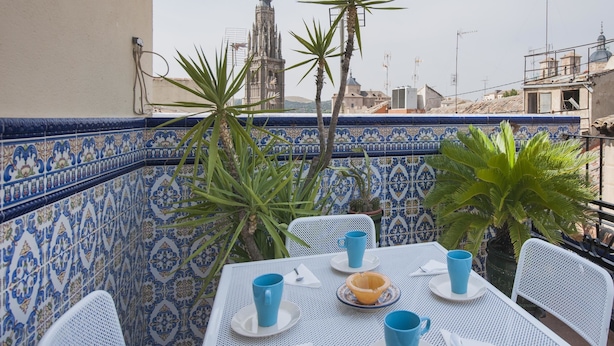 Taula preparada amb tasses blaves i un bol, envoltada de caires blancs, amb una paret de rajoles i una vista d'un edifici alt al fons.