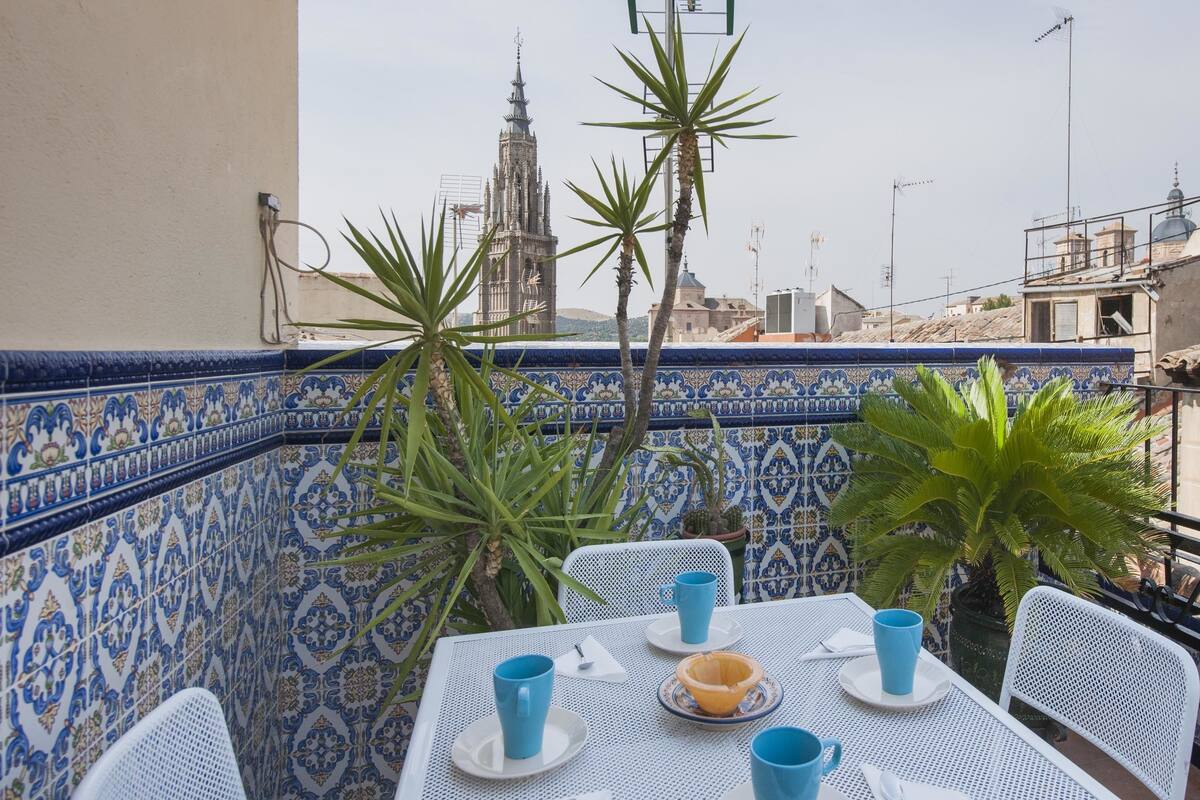 Una mesa dispuesta con tazas azules y un bol, rodeada de sillas blancas, con una pared de azulejos y una vista de un edificio alto al fondo.