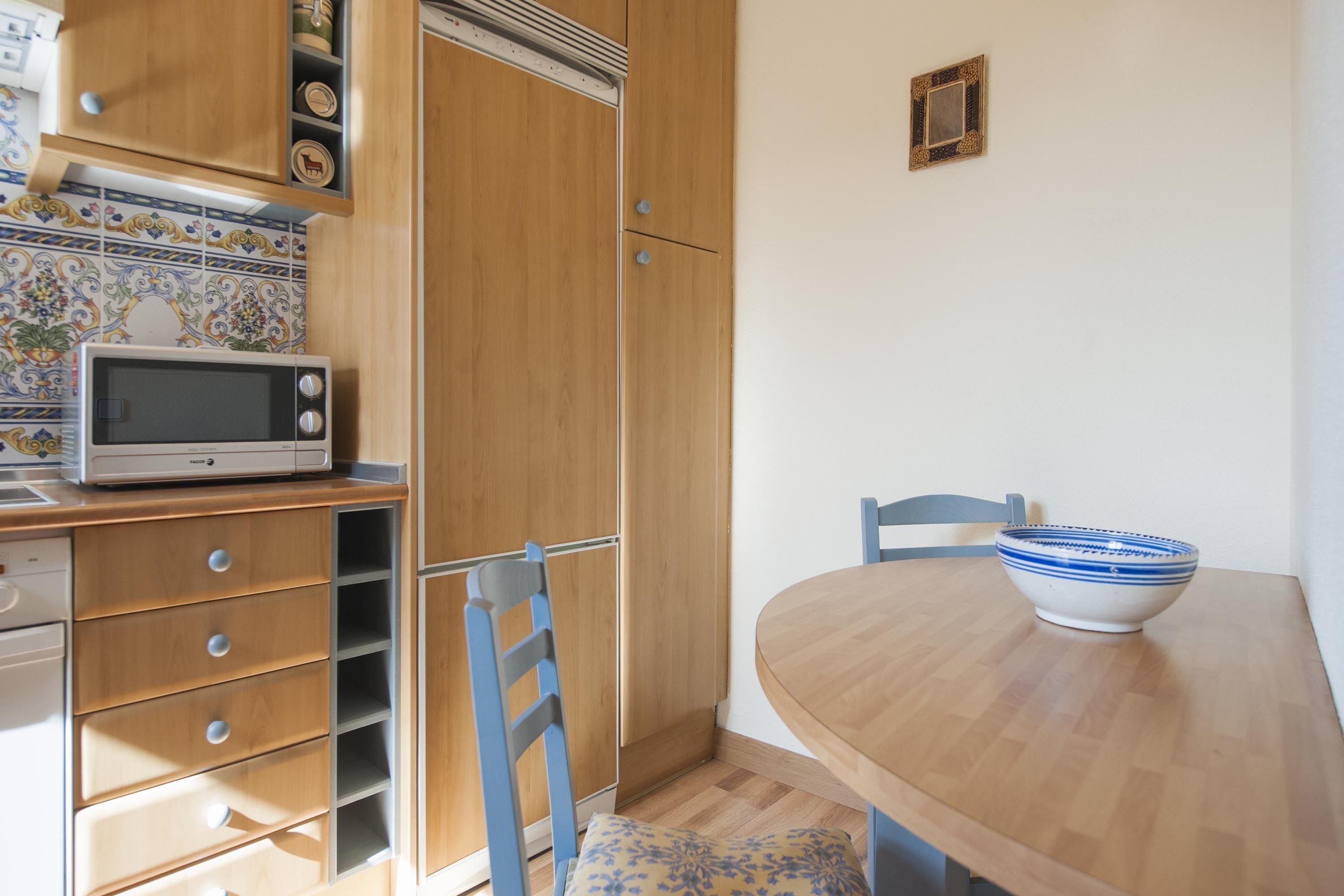 A compact kitchen with a microwave, wooden cabinets, and a dining table with a bowl.
