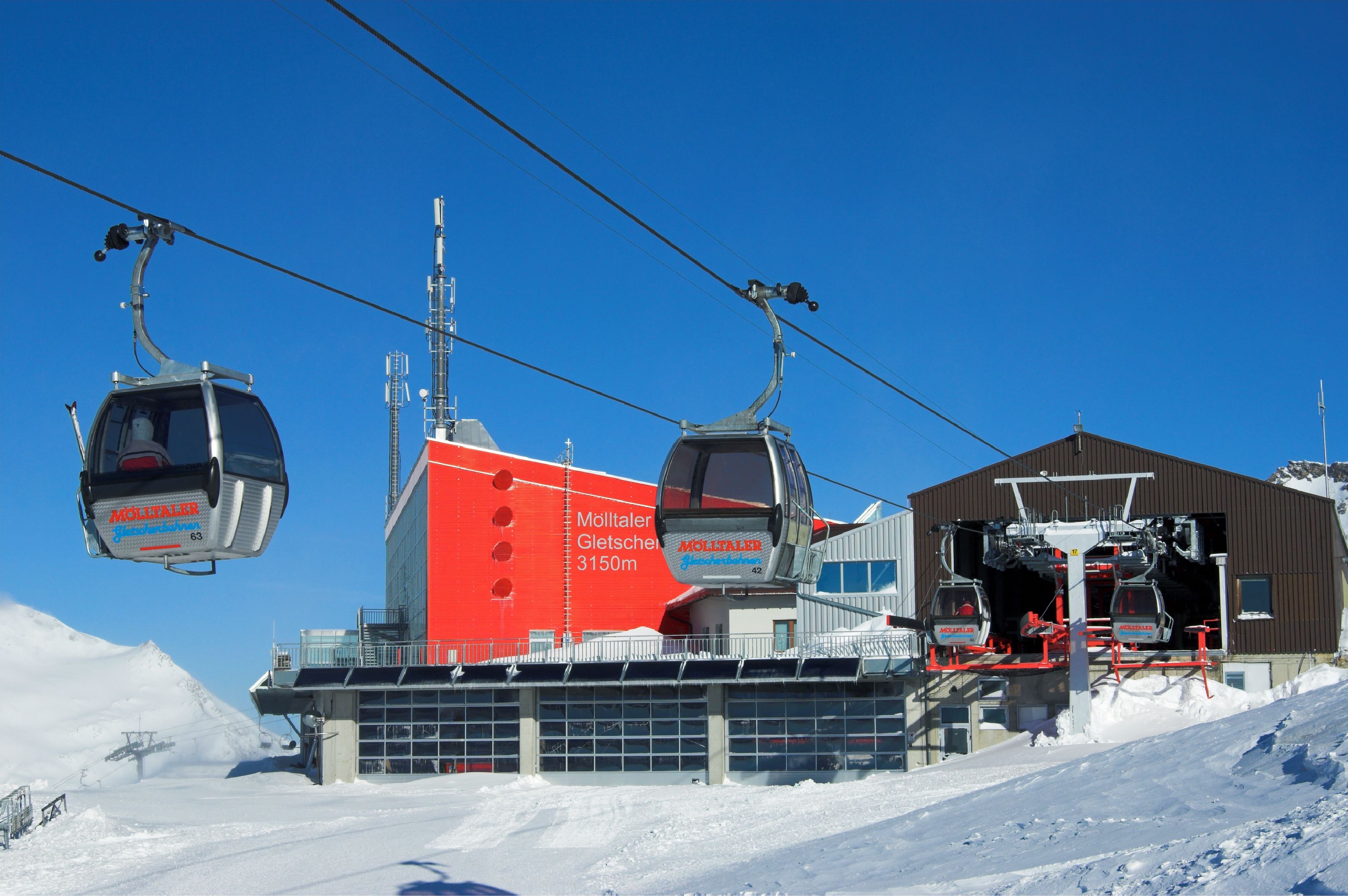 A ski lift with gondolas, a red building with 'Mölltal' signage, and a snow-covered slope.