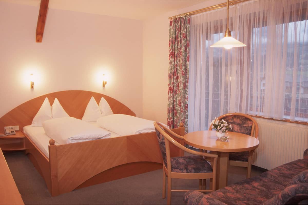 A hotel room with a wooden bed, a round dining table with a chair, a sofa, and a window with curtains.