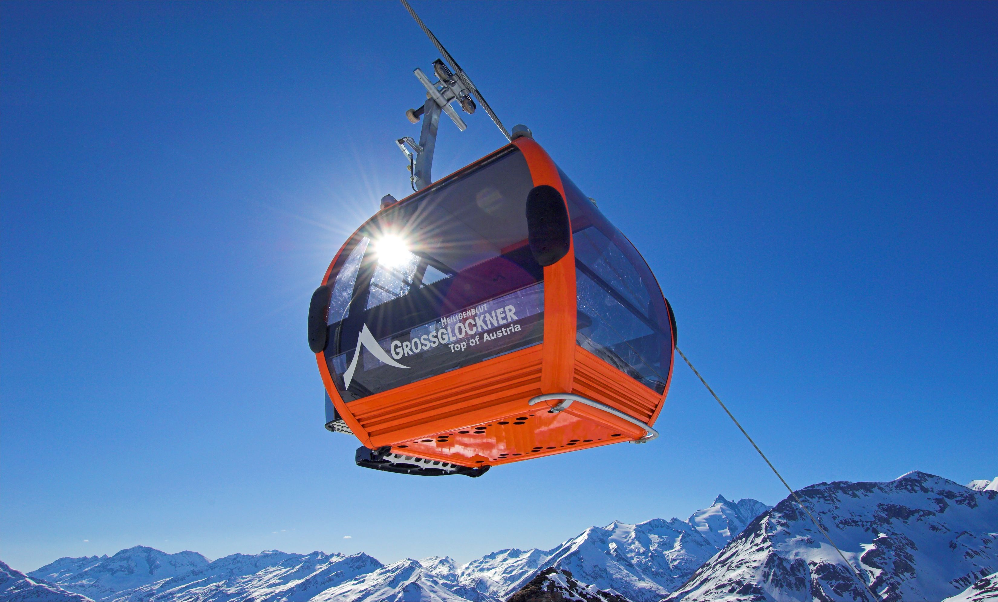 A cable car with "GROSSGLOCKNER Top of Austria" written on it, suspended by cables against a clear blue sky and snow-covered mountains.