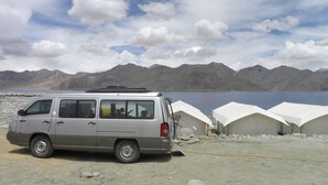 Una camioneta plateada estacionada cerca de tiendas de campaña blancas a orillas de un lago, con montañas al fondo.