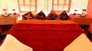Habitación de hotel con una cama grande, almohadas estampadas con motivos florales, una colcha roja y dos mesitas de noche con lámparas.