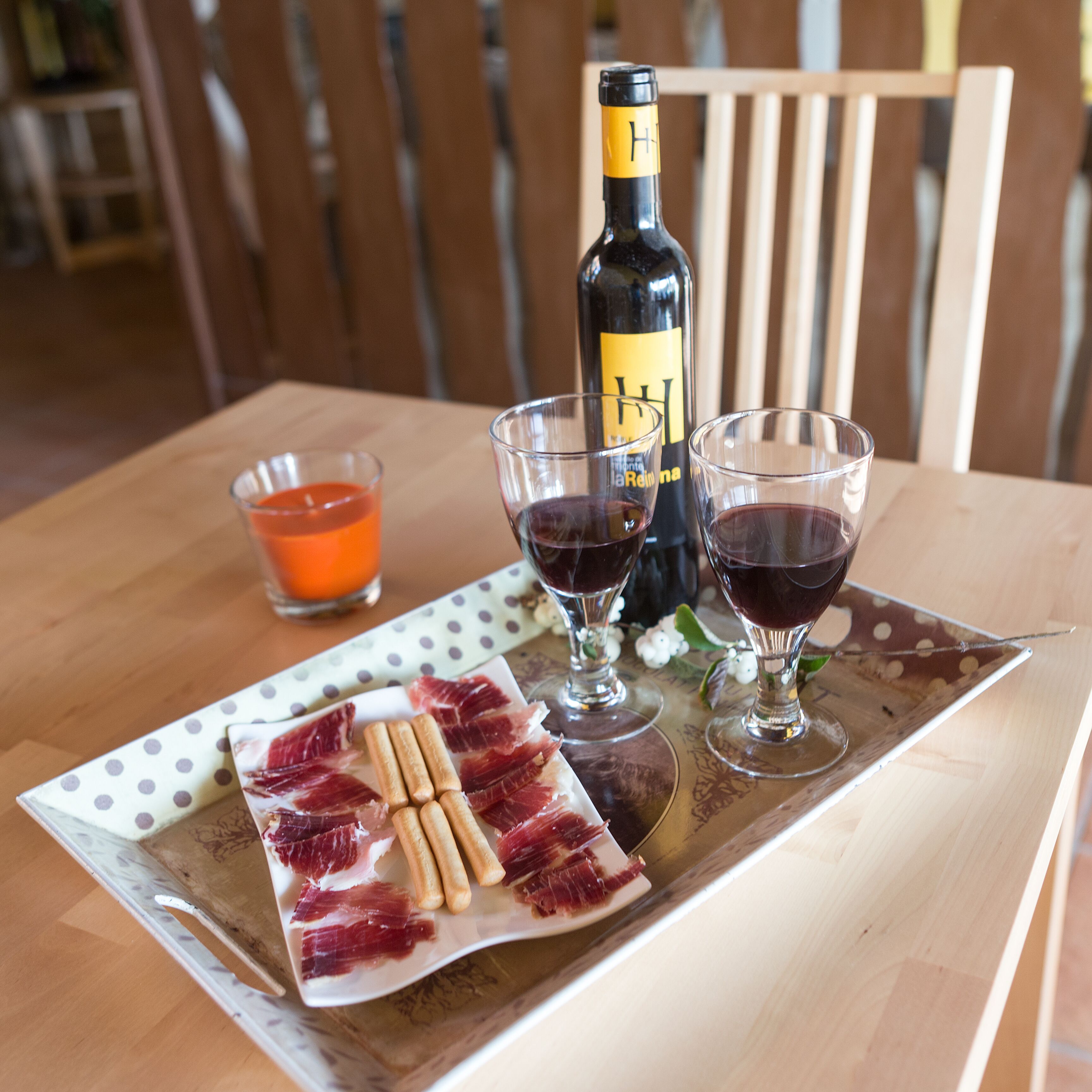 Una mesa de madera con una botella de vino, dos copas de vino tinto, un cuenco pequeño con líquido anaranjado, y un plato con lonchas de embutido y galletas.
