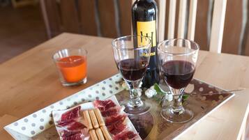 Una mesa de madera con una botella de vino, dos copas de vino tinto, un cuenco pequeño con líquido anaranjado, y un plato con lonchas de embutido y galletas.