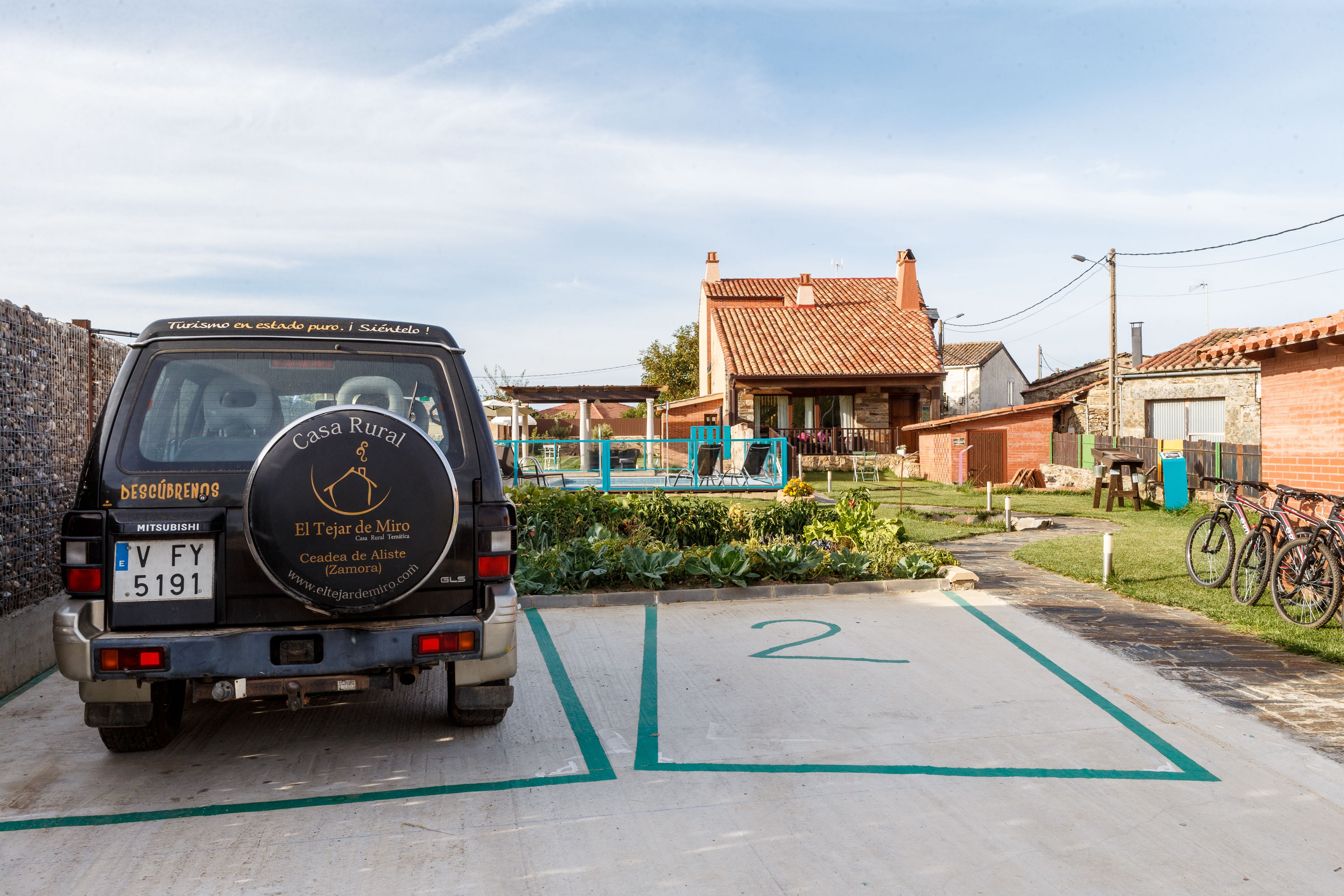 Casa rural con techo de tejas, zona ajardinada y un vehículo estacionado con matrícula visible.