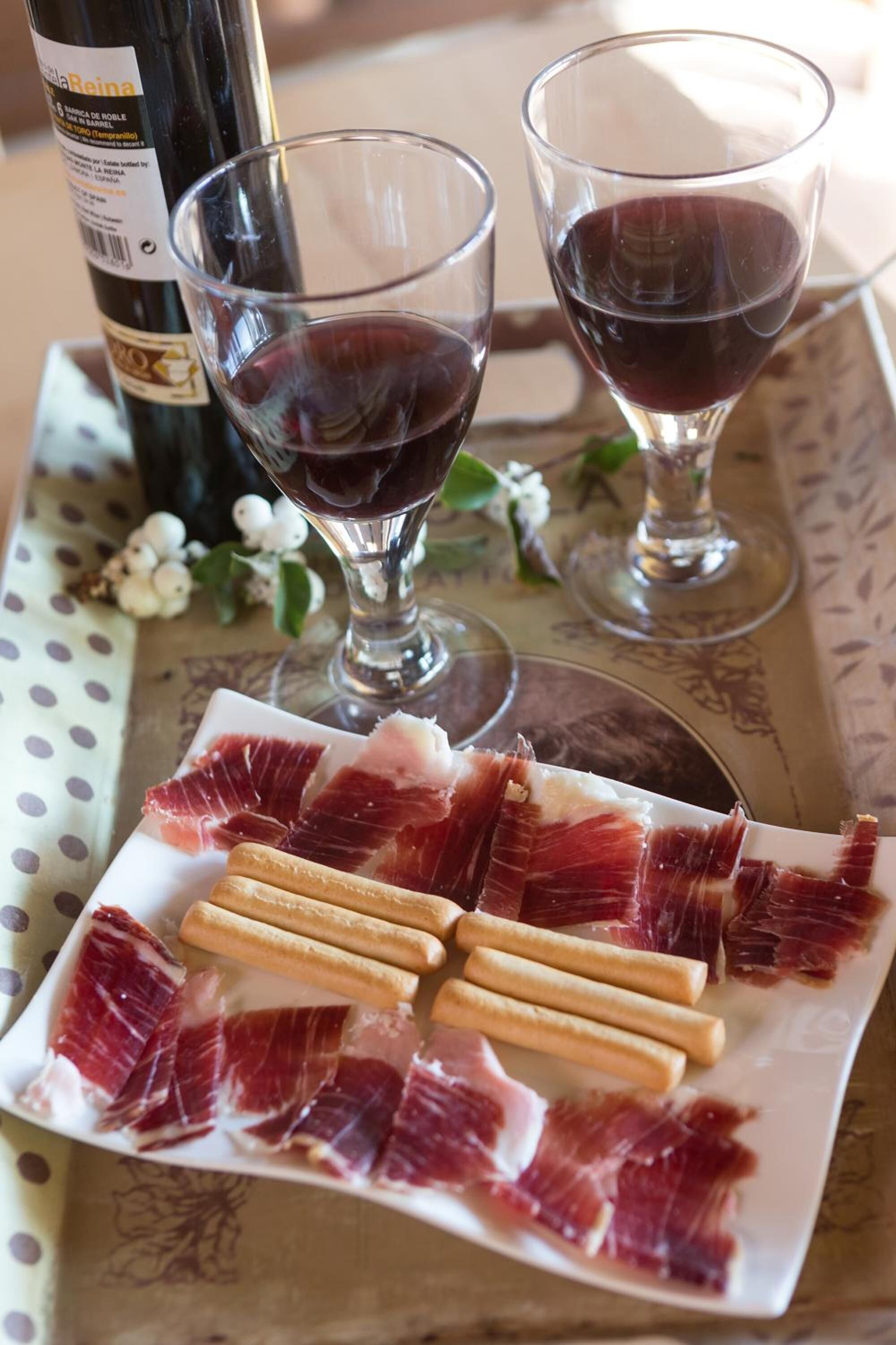 A plate of cured meat, a bottle of wine, and two glasses of red wine on a table with a polka dot tablecloth.
