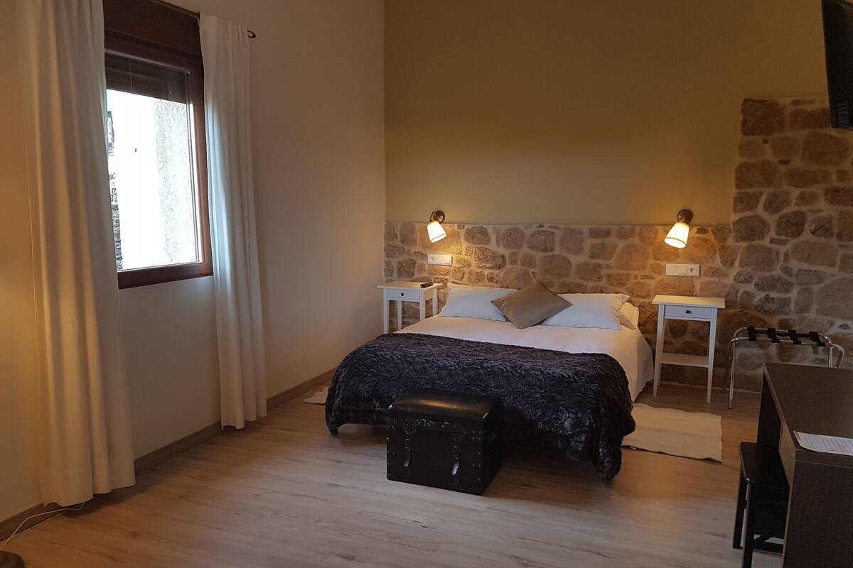 A bedroom with a stone wall, a bed with a dark patterned bedspread, two bedside lamps, a small wooden bench, and a desk with a chair.