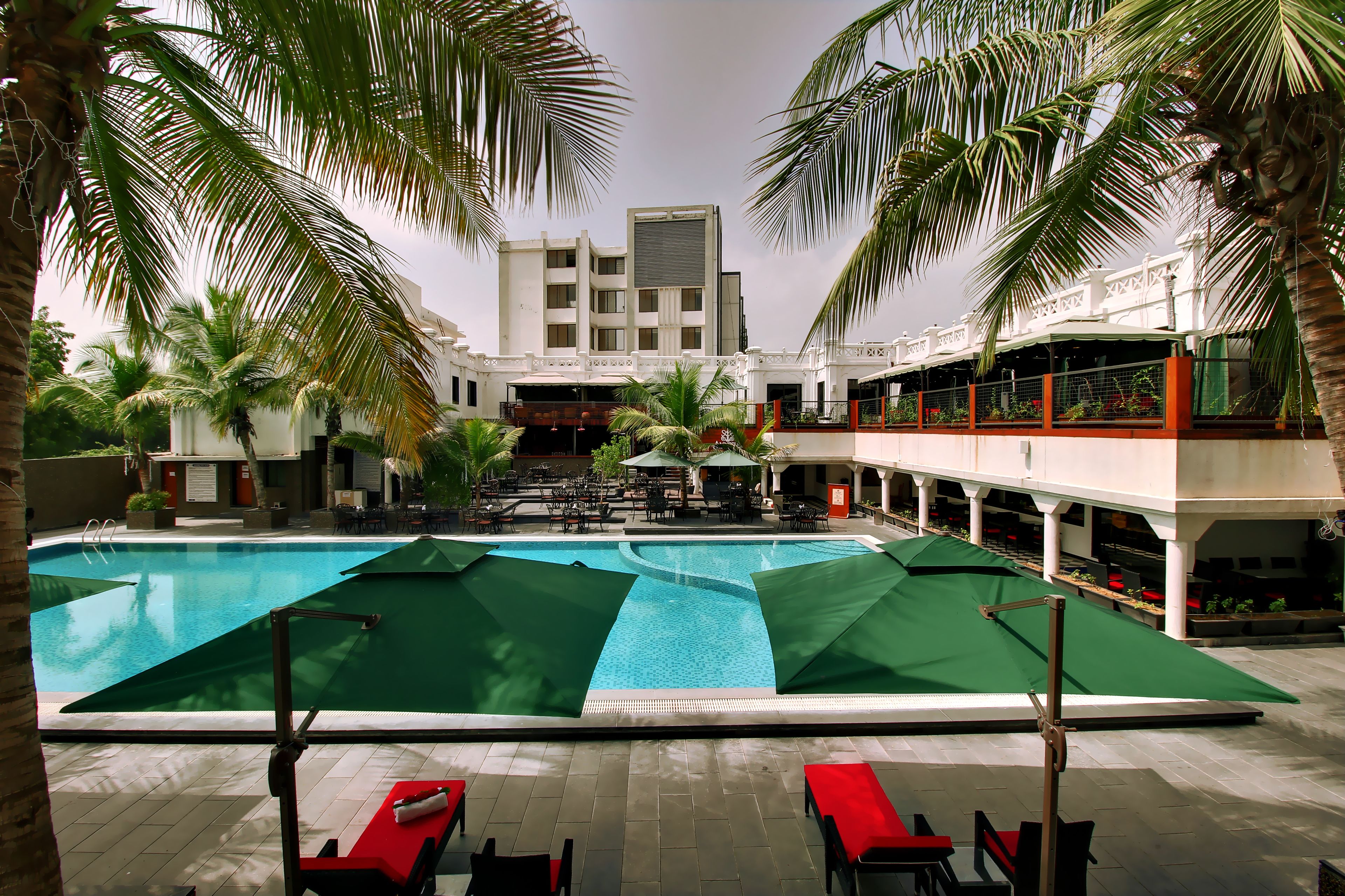 A swimming pool with lounge chairs and umbrellas, surrounded by palm trees and a building with balconies.