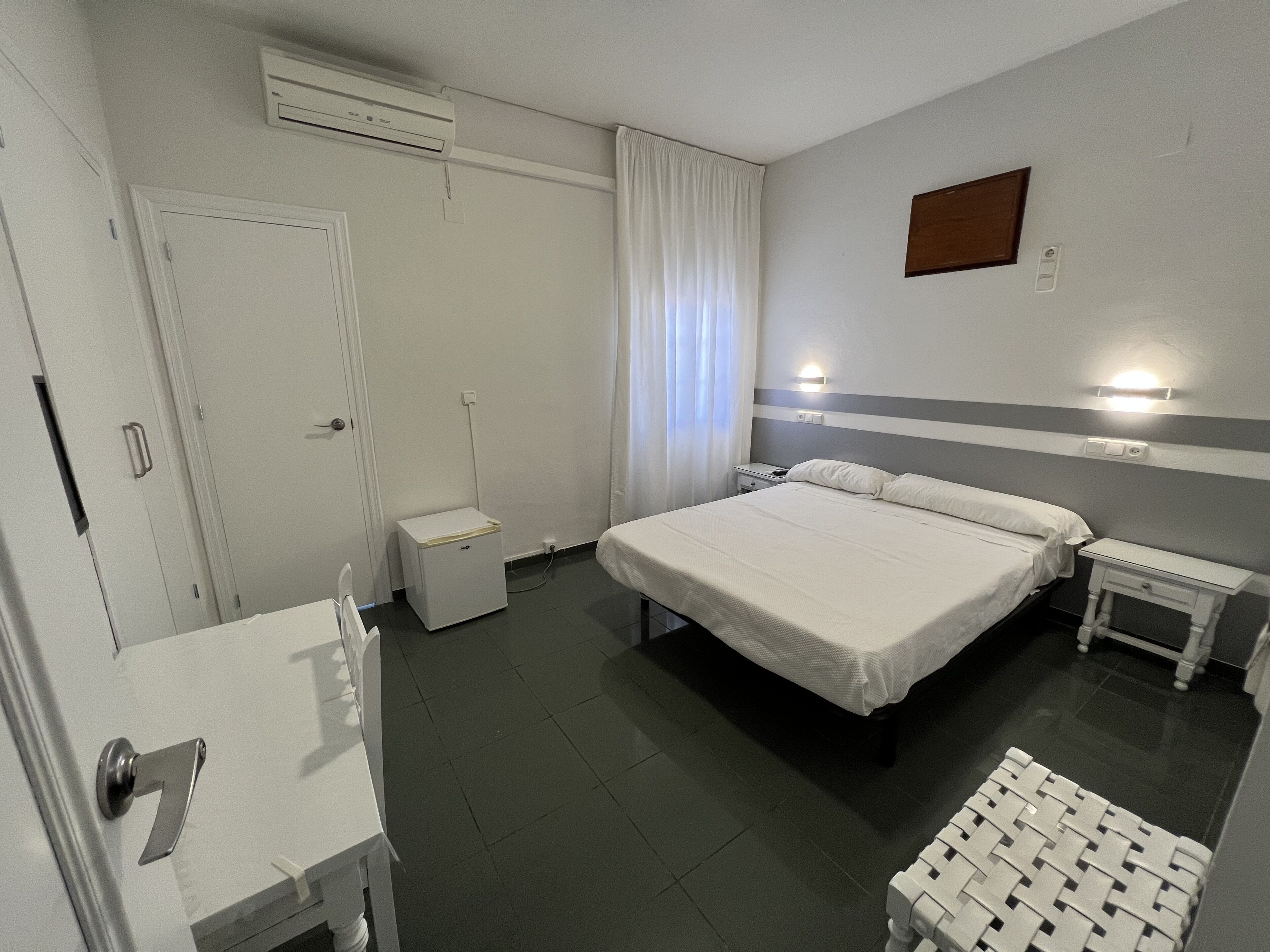 A hotel room with a bed, bedside tables, a desk, a chair, a wardrobe, and an air conditioning unit.