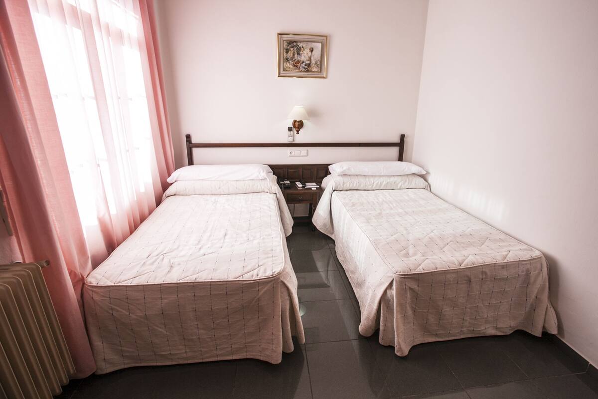 A hotel room with two single beds, a nightstand with a lamp, a framed picture on the wall, and a radiator.