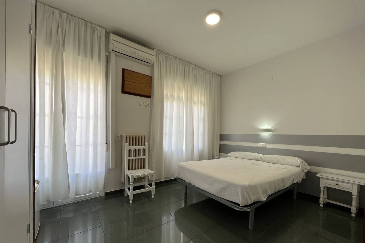 A hotel room with a bed, bedside tables, a radiator, and a window with curtains.