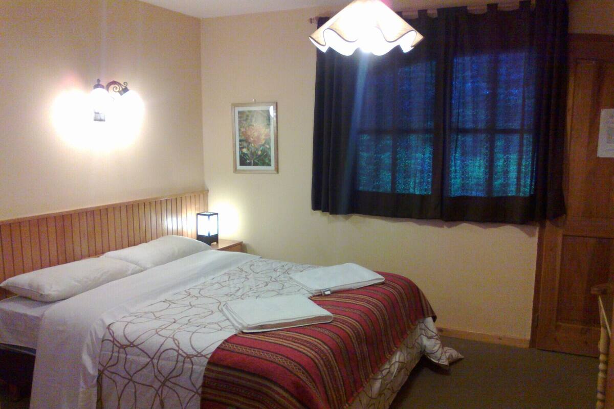 Una habitación de hotel con cama, mesita de noche, lámpara, un cuadro y una ventana con cortinas.