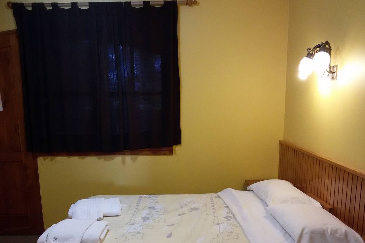 Una cama bien tendida con una colcha estampada con motivos florales, una cortina oscura y una lámpara fijada a la pared.
