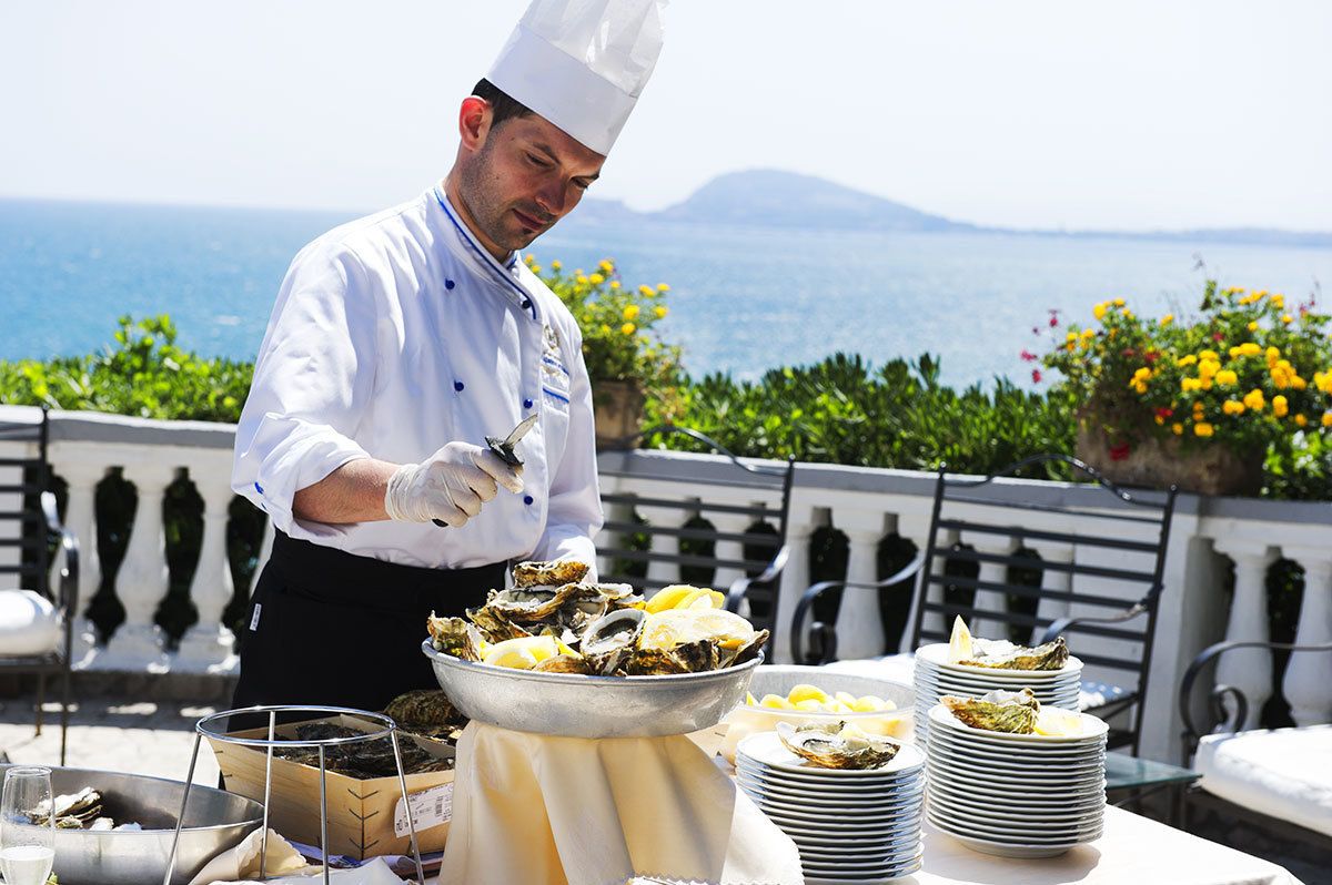 Un chef prépare des huîtres avec des quartiers de citron sur une terrasse offrant une vue sur la mer.