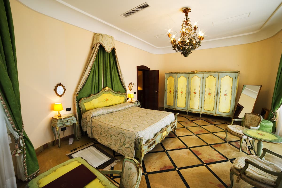 A bedroom with a four-poster bed, a chandelier, a green canopy, a vintage wardrobe, and a small table with a chair.