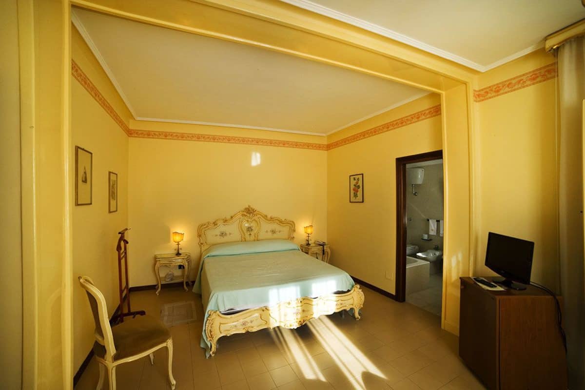 A hotel room with a large bed, bedside lamps, a chair, a television, and a bathroom visible through an open door.