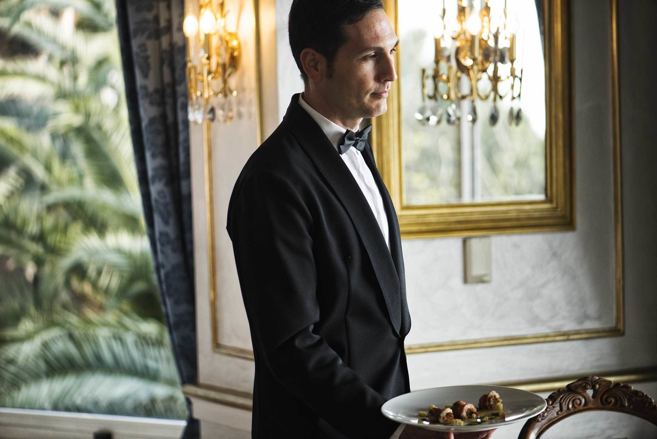 A man in a black tuxedo standing in a room with a large window, a chandelier, and a plate of food on a table.