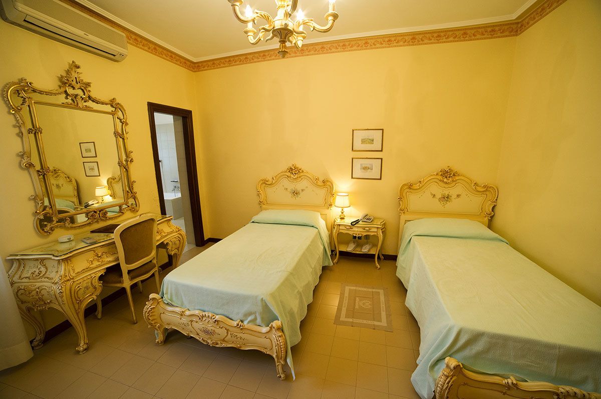 A hotel room with two beds, a desk with a chair, a large ornate mirror, and a chandelier.