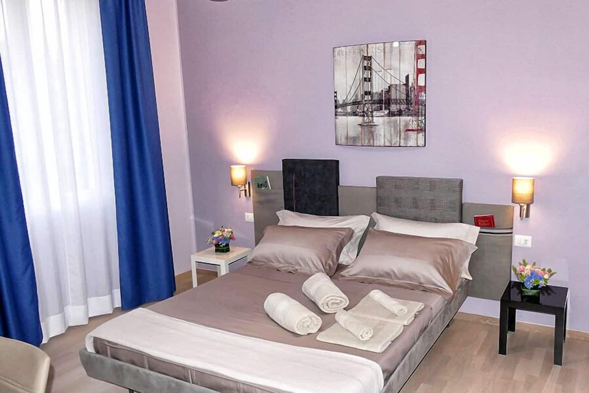 Ein Hotelzimmer mit Bett, zwei Nachttischen, einer Lampe, einem Bild an der Wand und einem Fenster mit Vorhängen.