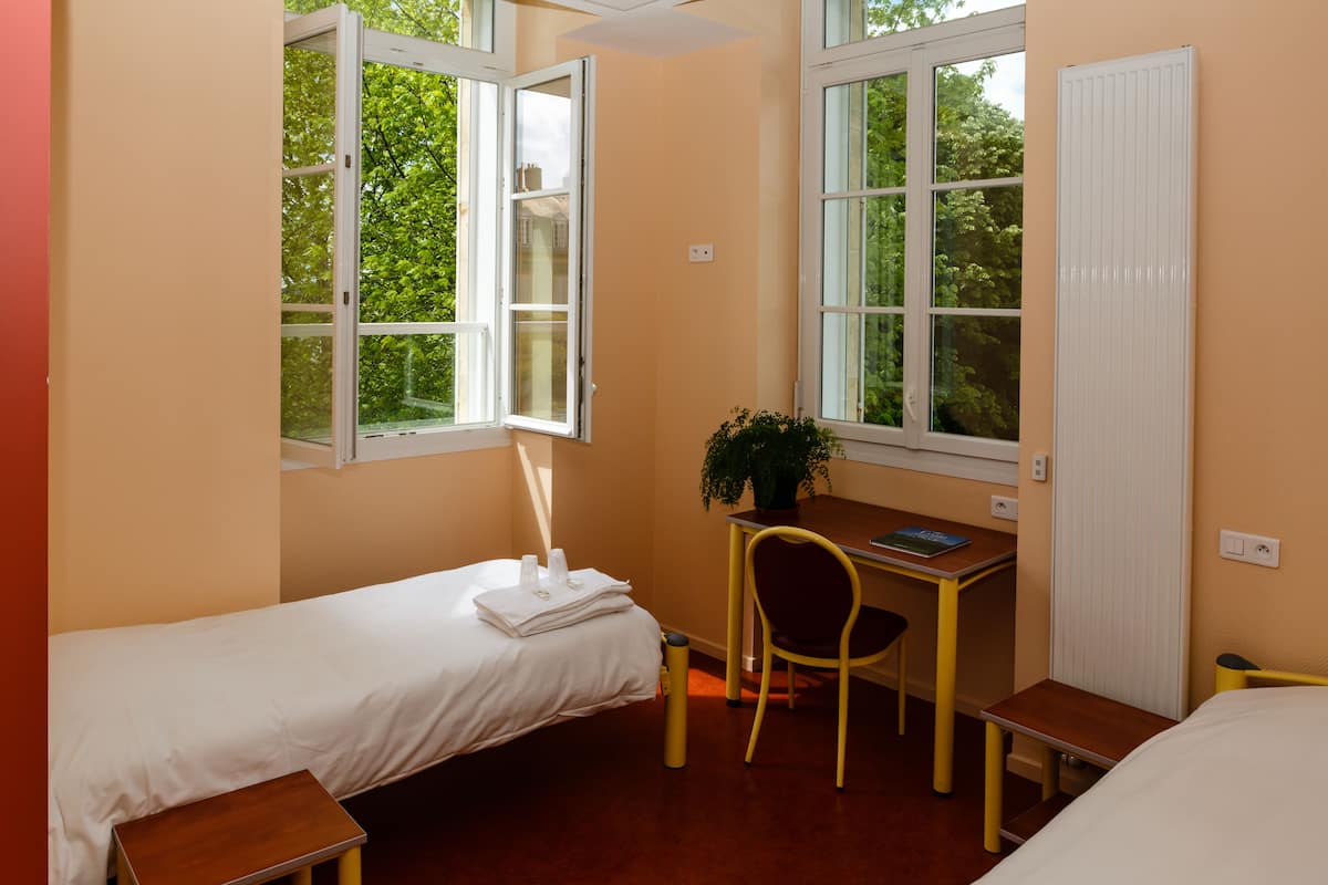 Una habitación pequeña y bien iluminada con una cama individual, un escritorio y una silla. Hay una ventana que deja entrar luz natural y ofrece vistas de la vegetación.