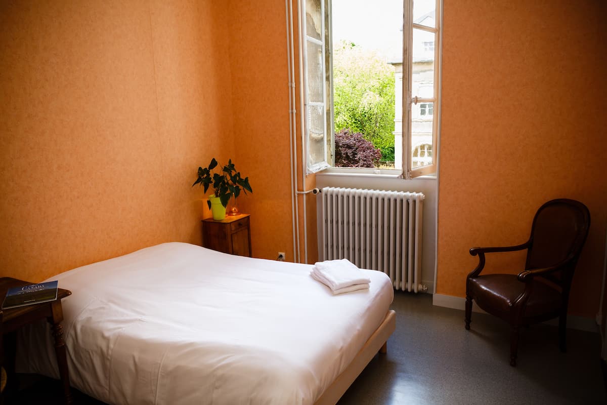Un dormitorio con cama, una silla, un radiador, una ventana y una pequeña mesa con una planta.