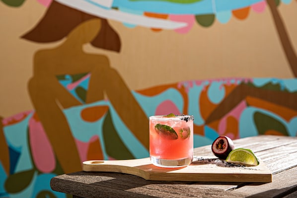 A glass of pink drink with lime on a wooden board, with a colorful mural in the background.