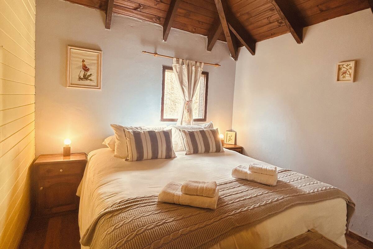 Um quarto aconchegante com teto de madeira, uma cama com travesseiros listrados, uma mesa de cabeceira com abajur e um banquinho de madeira.