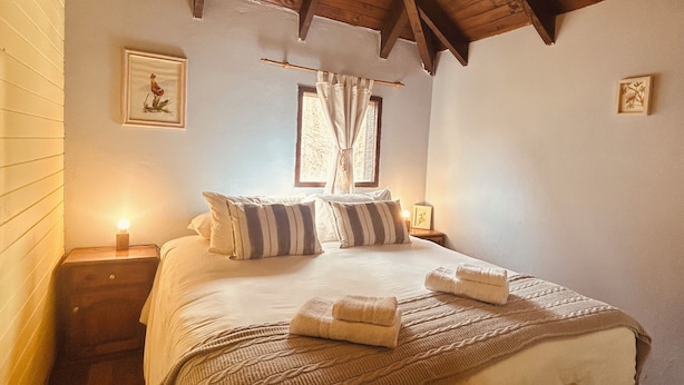Une chambre confortable avec un plafond en bois, un lit agrémenté d’oreillers à rayures, une table de chevet avec une lampe et un petit tabouret en bois.