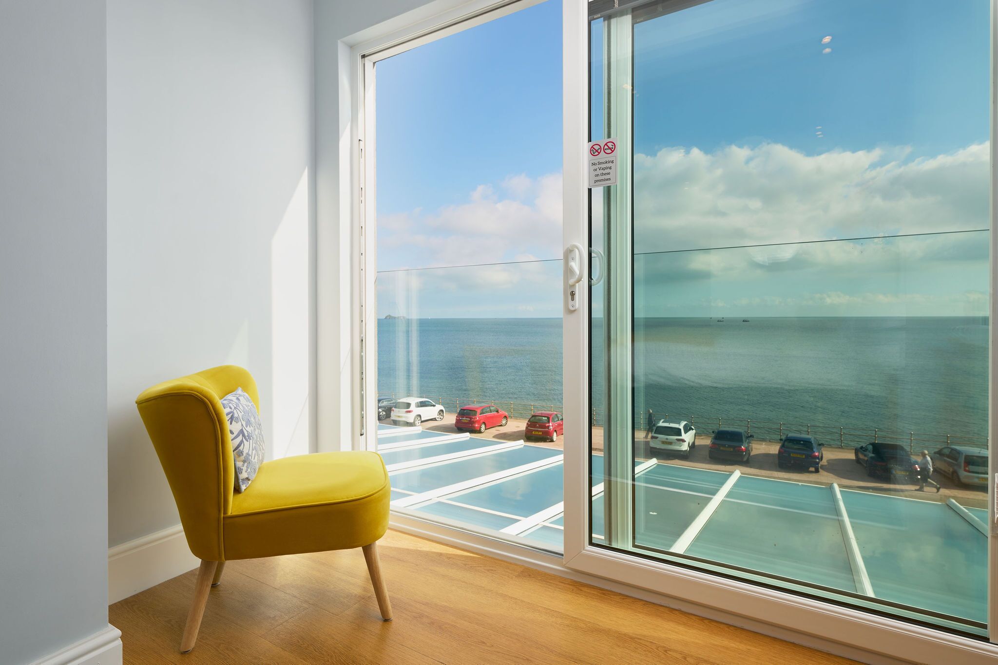 A room with a yellow chair, a large sliding glass door, and a view of the sea and parked cars.