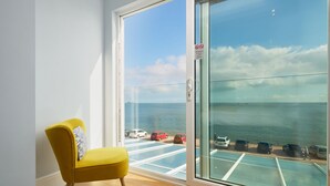A room with a yellow chair, a large sliding glass door, and a view of the sea and parked cars. - Channel View Hotel (Paignton)