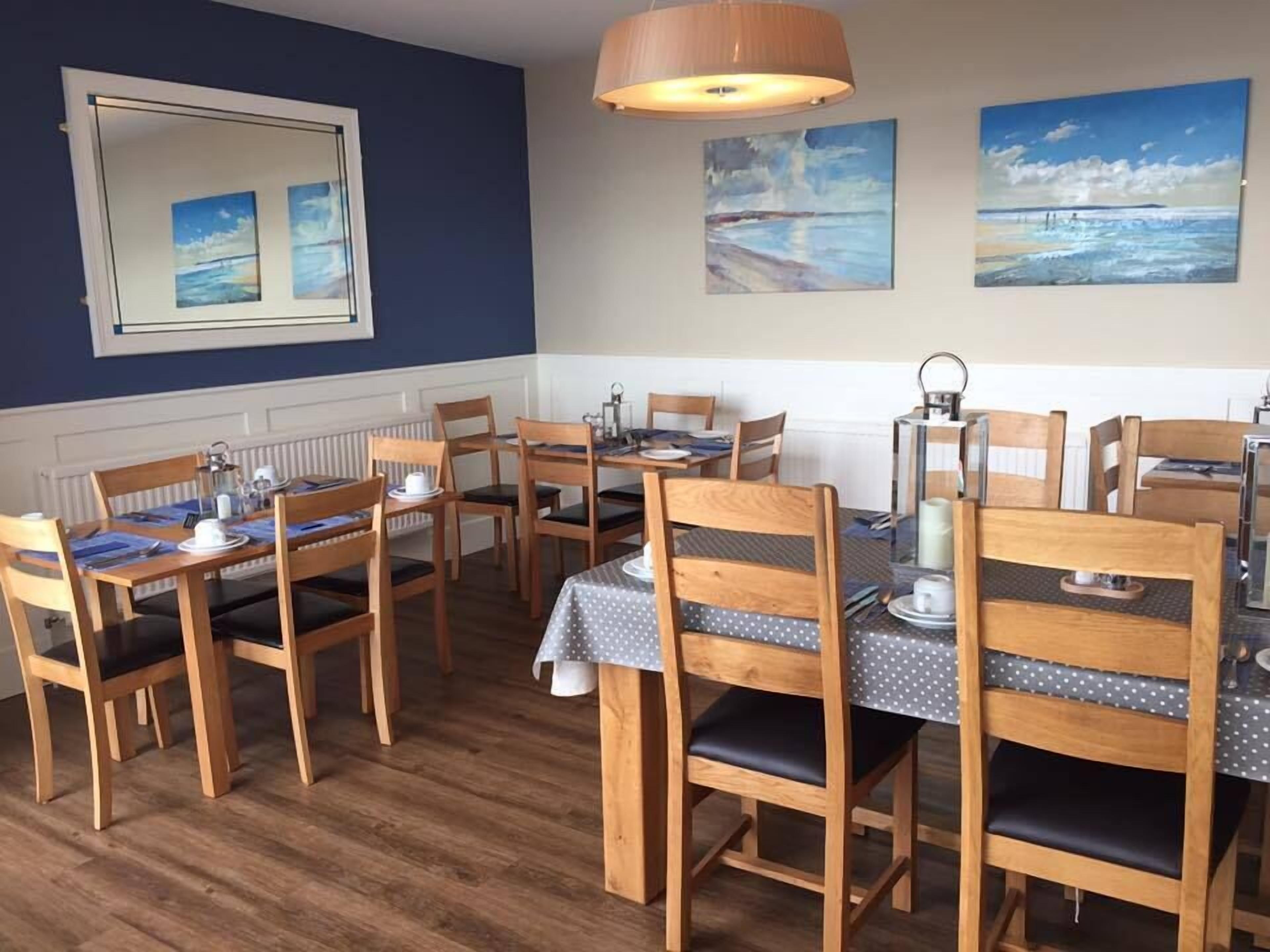 A dining area with wooden chairs and tables, a mirror on the wall, and paintings depicting coastal scenes.