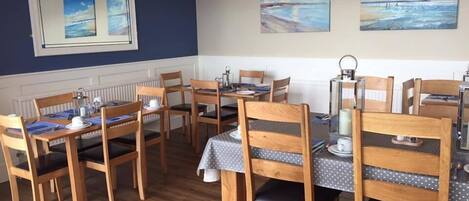 A dining area with wooden chairs and tables, a mirror on the wall, and paintings depicting coastal scenes.