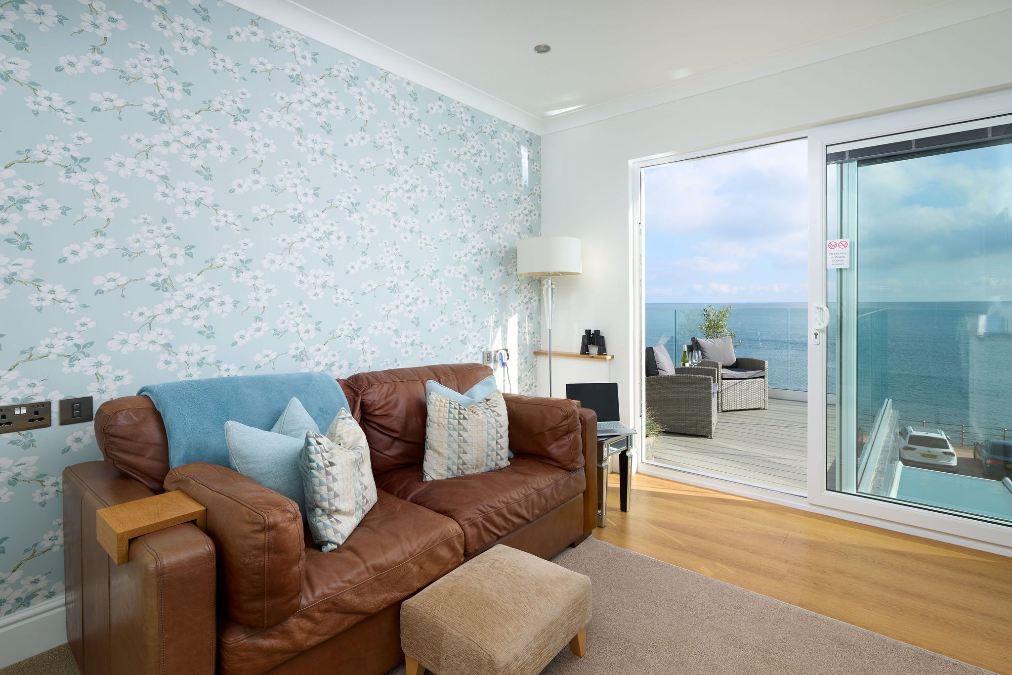 A modern living room with a brown leather sofa, a blue floral wallpaper, and a view of the sea through a glass door.