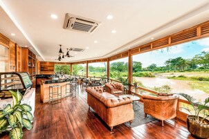 Amplia habitación con piso de madera, barra y grandes ventanas que dan a una vista del río y follaje.