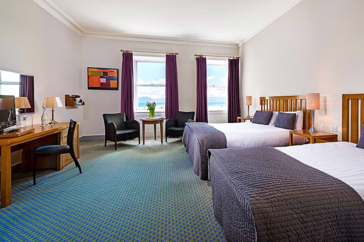 Une chambre d’hôtel avec deux lits, un bureau avec un ordinateur, une chaise, une petite table avec un vase et une fenêtre avec des rideaux.