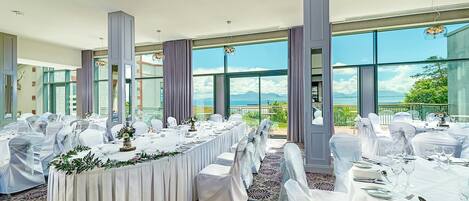 Un salón de banquetes con mesas redondas dispuestas para un evento formal, ventanas amplias que ofrecen vistas panorámicas y un techo alto con iluminación empotrada.