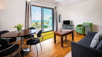 A modern living room with a dining area, a green sofa, a TV, and a balcony with a view of the sea.