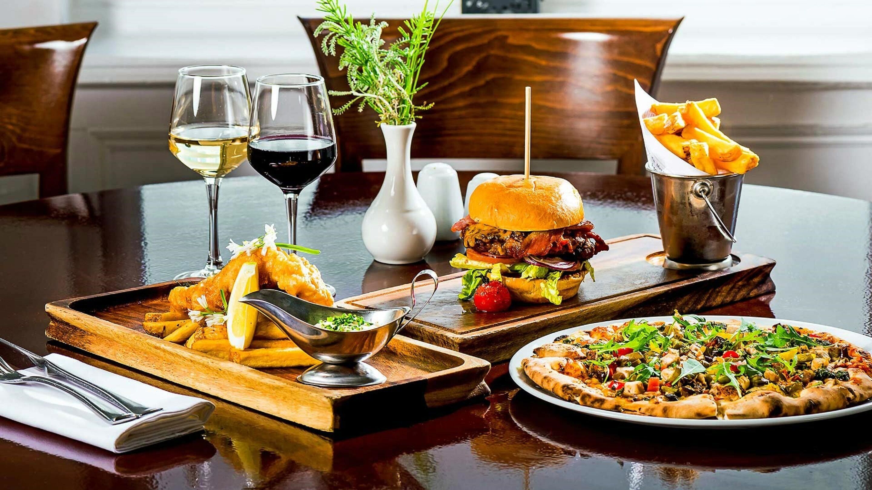 A dining table with a variety of dishes including fish and chips, a burger, a pizza, and a steak. There are also two glasses of wine and a vase with greenery.