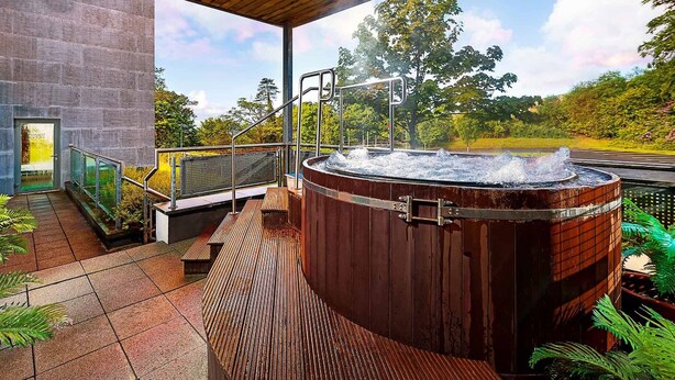 A hot tub with a wooden exterior, a metal railing, and a view of a road and greenery. - Mulranny Park Hotel (Mulranny)