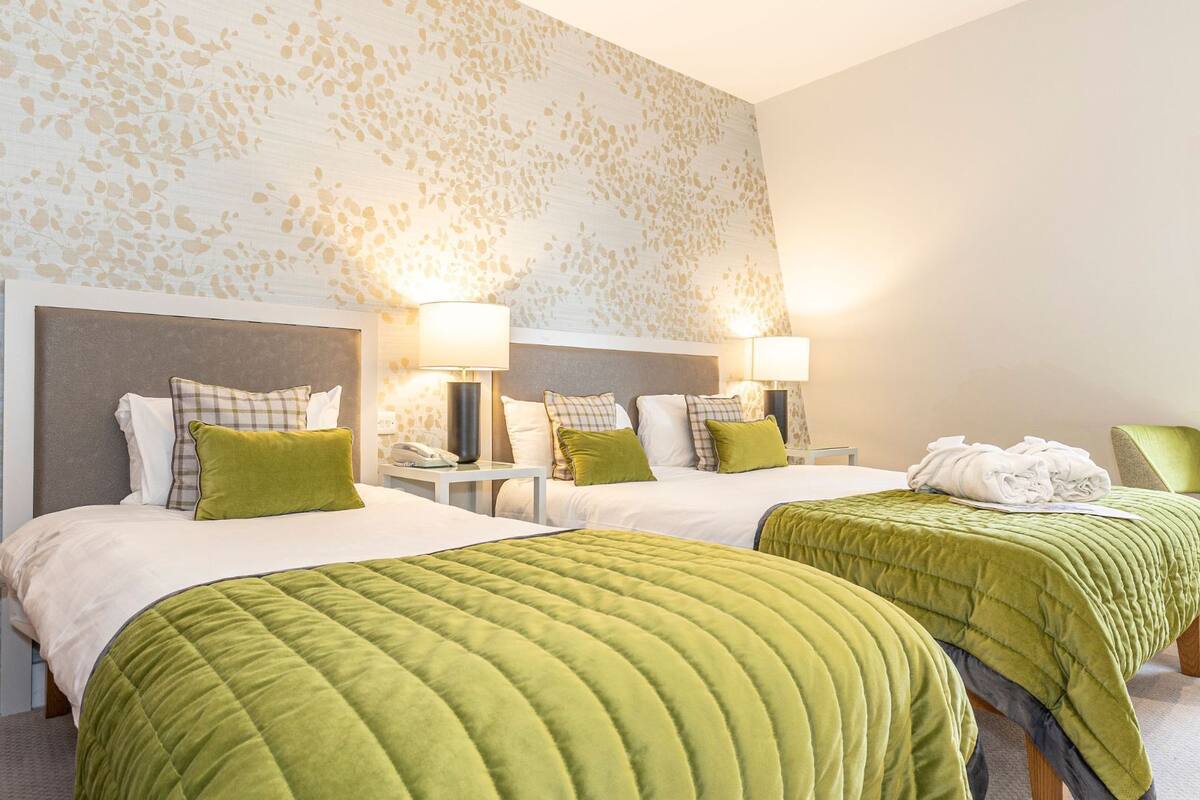 Une chambre d’hôtel avec deux lits, chacun recouvert d’une courtepointe matelassée verte et agrémenté d’oreillers assortis. Il y a des tables de chevet avec des lampes, et une serviette pliée est posée sur la table de droite.