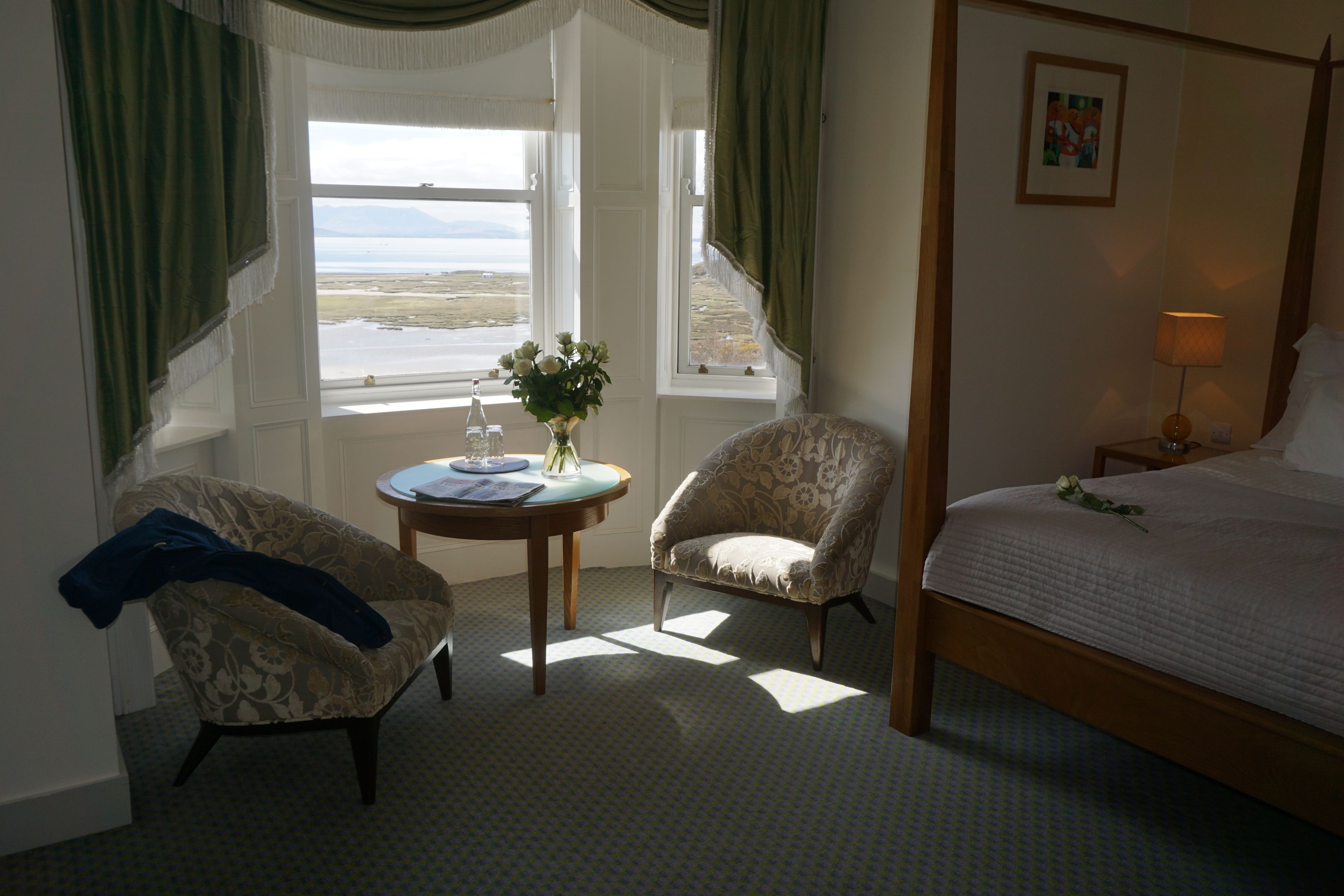 A bedroom with a four-poster bed, a chair, a small table with a vase of flowers, and a window with a view of the sea.